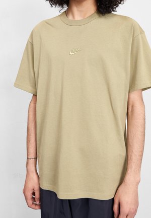 Personne portant un t-shirt Nike ample de couleur marron clair avec un petit logo sur la poitrine et un pantalon foncé, debout devant un fond uni.