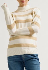 Gestreifter Strickpullover in Creme und Beige, mit hohem Kragen, gerippten Bündchen und strukturiertem Stoff. Kombiniert mit blauen Jeans.