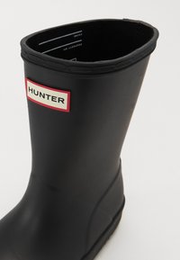 Botte de pluie en caoutchouc noir avec un bord supérieur renforcé, patch logo rouge et blanc, semelle texturée et finition lisse.