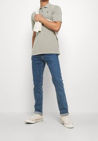 Šedé polo tričko s krátkými rukávy, modré džíny a bílé tenisky. Polo je jednoduché, s malým logem. Model drží lehký svetr.