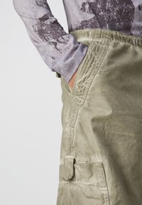 Olijfgroene cargoshorts van een gestructureerde stof, met zijzakken en zichtbare stiksels. Gecombineerd met een grijze top met lange mouwen en een patroon.