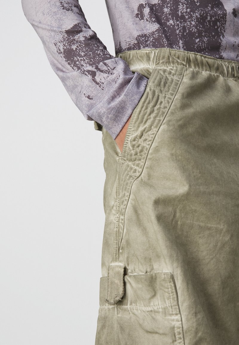 Olijfgroene cargoshorts van een gestructureerde stof, met zijzakken en zichtbare stiksels. Gecombineerd met een grijze top met lange mouwen en een patroon.