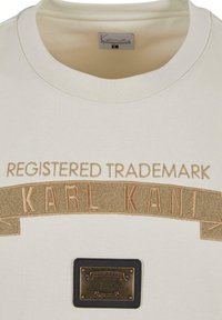 Kreemist puuvillast T-särk, millel on kuldsed tikitud tähed "REGISTREERITUD KAUBAMÄRK KARL KANI" ja metalline silt brändi logoga.
