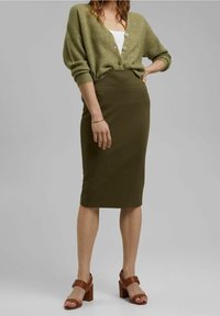Olijfgroen ribgebreid vest, aansluitende olijfgroene rok en bruine sandalen met blokhak. Gestepte brei en op maat gemaakte silhouet met knoopdetails.