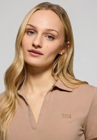 Krótki rękaw, beżowa koszulka polo z dekoltem w kształcie litery V. Zawiera subtelny, wyhaftowany logo "POLO CLUB" na piersi. Gładka tekstura materiału.