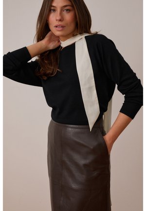 Kvinde iført sort langærmet top, mørkebrun lædernederdel og lys beige halsrør, med den ene hånd, der rører ved hendes hår og den anden i lommen.