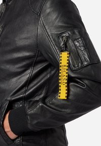 Zwarte leren jas met ribgebreide manchetten, ritszak op de mouw en een gele label met zwart patroon. Gladde textuur en aansluitend ontwerp.