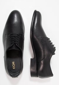 Paire de chaussures habillées noires en cuir à lacets, l'une montrant une vue de dessus avec la marque "KIOMI" à l'intérieur, l'autre montrant un profil latéral.