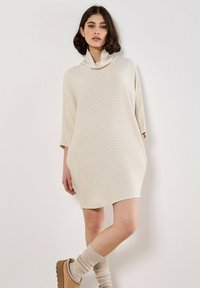 Abito maglione oversize di colore crema con collo alto, texture a coste e spacchi laterali. Abbinato a calze al ginocchio e stivaletti slip-on.