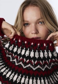 Pull en tricot avec un col haut, présentant un motif en zigzag noir, blanc et rouge. Accentué de bijoux en gemmes noires et transparentes.