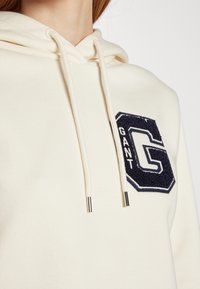 Sudadera blanca con capucha y cordones, con un parche texturizado en azul marino oscuro que lleva la letra "G" y la palabra "GANT" en blanco. Tela suave.