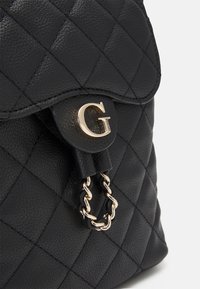 Mochila negra de cuero acolchado con un destacado hardware en tono dorado en forma de letra G y un acento de cadena adjunto al cierre.