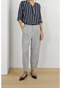Frau in einem hochgekrempelten navy gestreiften Bluse, hochtaillierten hellgrauen Hosen, schwarzem Gürtel und schwarzen spitzen Slingback-Heels, die drinnen steht.