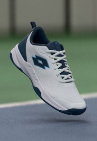 Weißes und marineblaues Sportschuh mit Schnürsenkeln und einem blauen geometrischen Logo, schwebend über einer Tennisspielfläche.