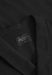 Crna bluzica s mekanom, glatkom teksturom. Ima V-izrez i oznaku "Vaschö Stockholm S." Jednostavan, elegantan dizajn.