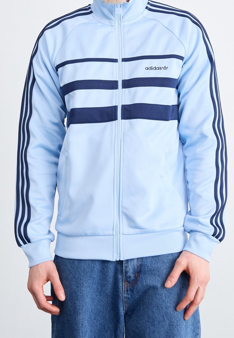 Muž v světle modré bundě Adidas s tmavě modrými pruhy a logem, v kombinaci s modrými džíny, s rukama u boků.