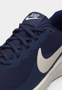 Zapatilla de running Nike azul marino con logo swoosh blanco, parte superior de malla y cordones azules atados sobre una lengüeta acolchada con la marca Nike.