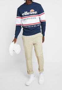 Man i marinblå, vit och röd ellesse-tröja, beiga uppvikta byxor, vita sneakers, håller en vit hatt i vänster hand.