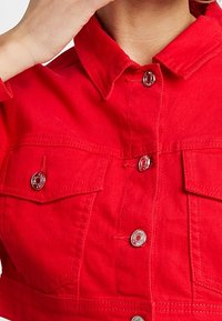 Červená zkrácená džínová bunda s límečkem, dvěma hrudními kapsami, stříbrnými patentkami a viditelnými švy.