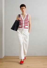 Rood-wit gestreepte mouwloze top met knopen, witte wijde pijpenbroek en rode schoenen. Zwarte totebag. Neutrale achtergrond en vloer.