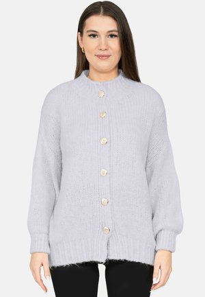 Vrouw met steil bruin haar, gekleed in een lichtgrijze gebreide cardigan met gouden knopen en een zwarte broek, staand tegen een witte achtergrond.