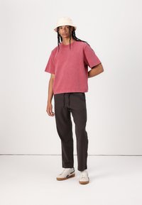 Rød bomulls oversized t-skjorte med korte ermer, kombinert med mørk grå bukser og beige bøttehatt. Hvite sneakers med brune såler synlige.