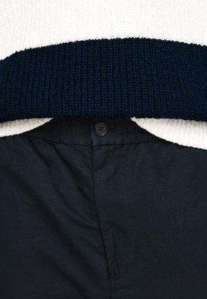 Gebreide trui met crème- en marineblauwe strepen, gedragen boven zwarte broeken. De trui heeft een gestructureerde geribbelde zoom, en de broek heeft een knoopsluiting.