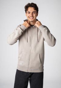 Lichtgrijze zip-up hoodie met drawstrings, zijzakken, ribbelmanchetten en een klein logo op de borst. Zacht stof met een gladde textuur.