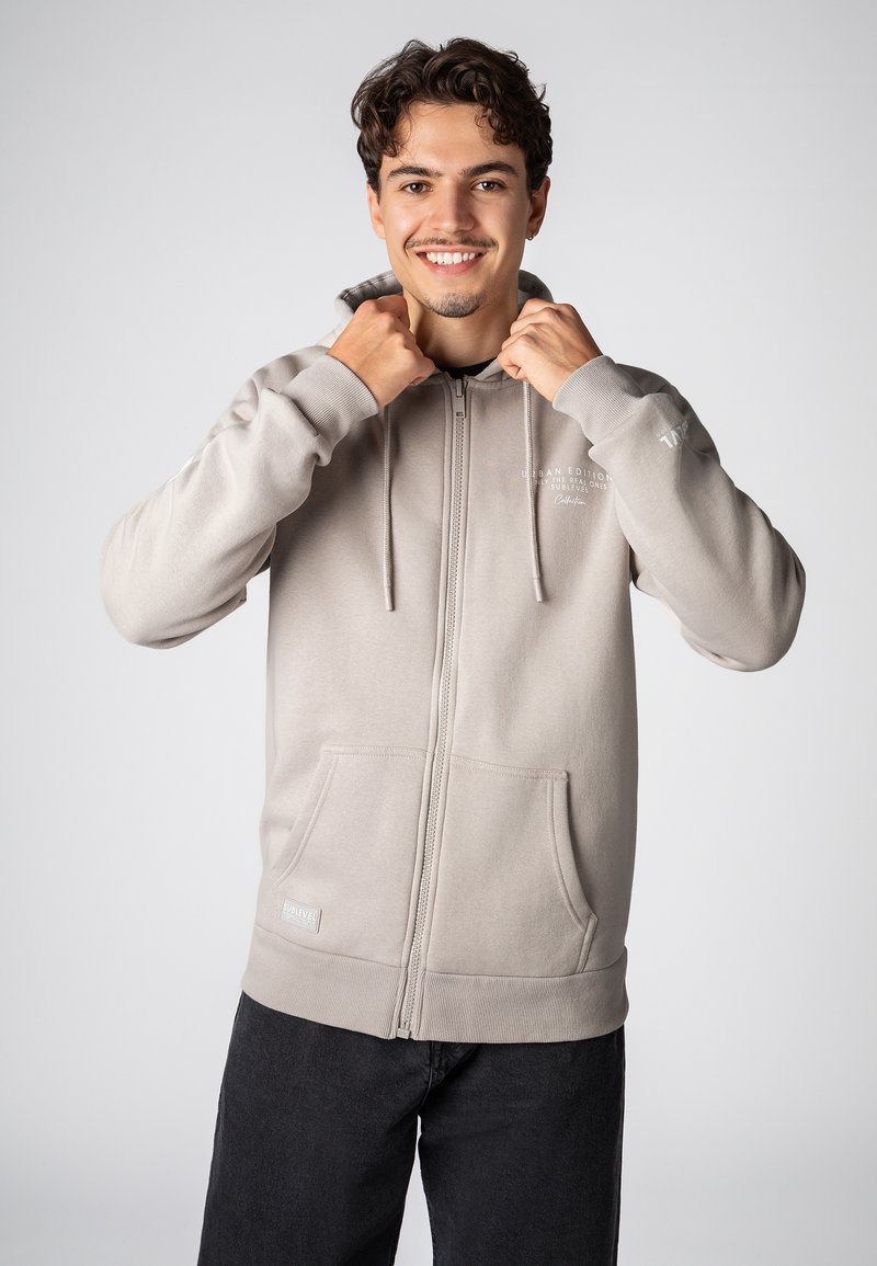 Lichtgrijze zip-up hoodie met drawstrings, zijzakken, ribbelmanchetten en een klein logo op de borst. Zacht stof met een gladde textuur.