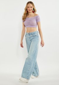 Jasnofioletowy, koronkowy crop top bez rękawów w połączeniu z wysokim stanem, jasnoniebieskimi, szerokimi dżinsami i białymi trampkami. Modelka idzie.