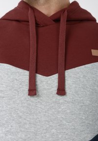 Μακρυμάνικο φούτερ με κουκούλα σε μπορντό και γκρι χρώμα, με μοτίβο ψαροκόκκαλο, που διαθέτει δύο μπορντό κορδόνια και μια μικρή ταμπέλα σε ανοιχτό καφέ χρώμα στο πλάι.