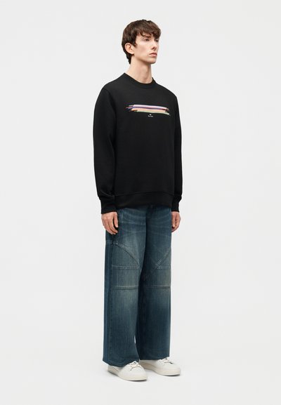 Νέος άνδρας που στέκεται, φοράει μαύρο φούτερ με πολύχρωμες ρίγες, φαρδιά μπλε τζιν παντελόνια και λευκά αθλητικά σε απλό λευκό φόντο.