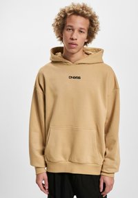 Sudadera beige con capucha, bolsillo frontal y logo negro. Tejido suave, corte oversize, puños y dobladillo de canalé. Diseño liso sin patrones.