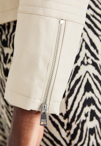 Manga de chaqueta de cuero crema con detalle de cremallera plateada. Costuras texturizadas cerca del hombro, con un fondo de patrón en blanco y negro en contraste.