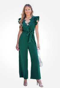 Groene jumpsuit met een kanten lijfje, ruches op de mouwen en een strikceintuur. Gecombineerd met heldere hakken en een lichte handtas.
