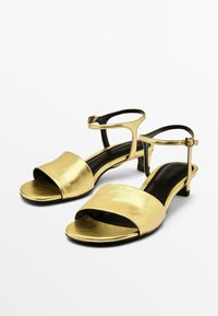 Sandales métalliques dorées avec un talon plat, bout ouvert et bride de cheville. Texture lisse avec une large bande avant et un intérieur noir.