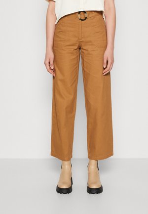 Frau trägt braune, hochtaillierte, weit geschnittene Hose mit Fronttaschen und Gürtel, kombiniert mit beigen Stiefeletten mit klobiger Sohle.