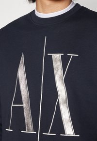 Felpa blu scuro con logo "A/X" ricamato in argento, scollatura rotonda e tessuto testurizzato. Primo piano sui dettagli delle lettere.