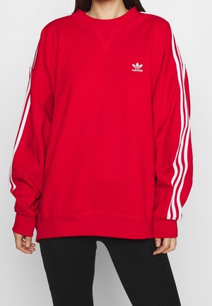 Sweatshirt rouge avec un col rond, des manches longues, logo Adidas blanc sur la poitrine et bandes blanches le long des manches. Texture de tissu douce.