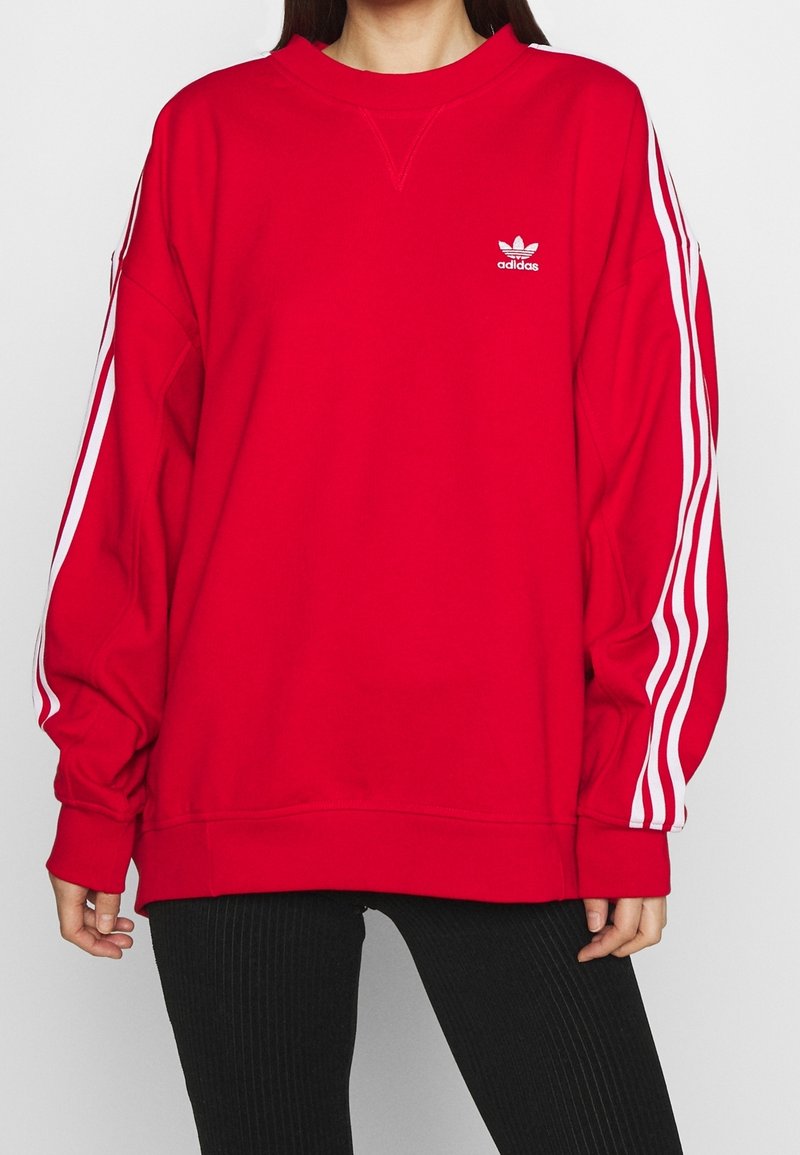 Röd sweatshirt med rund hals, långa ärmar, vit Adidas-logotyp på bröstet och vita ränder längs ärmarna. Mjuk tygtextur.