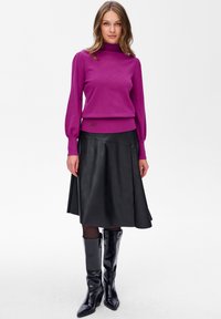 Kobieta w fioletowym swetrze z długim rękawem, czarnej skórzanej spódnicy w kształcie litery A, czarnych rajstopach i czarnych kozakach za kolano stoi na białym tle.