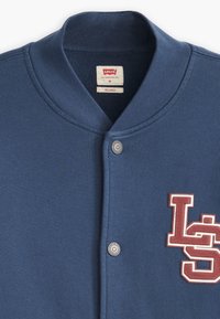 Cardigan azul estilo béisbol con un cuello acanalado, botón de presión con marca y un logo texturizado rojo "LS" en el frente. Fabricado con tela suave.