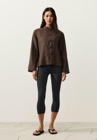 Vrouw draagt een bruin langermig topje met knopen, zwarte cropped leggings en zwarte teenslippers, staand op een stenen vloer tegen een effen muur.