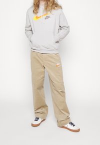 Grå hoodie med orange och svart Nike-logotyp, kombinerad med khakifärgade manchesterbyxor med Nike-märke. Vita sneakers med bruna sulor.