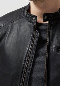 Svart läderjacka med dragkedjestängning och tryckknappskrage, med texturerad detaljering och slät finish, bärs över en mörkgrå tröja.