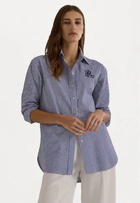 Svītraini pogu krekls tumši zils un balts, ar klasisku apkakli un mazu logotipu uz kreisā krūšu daļas. Izgatavots no gluda, viegla auduma.