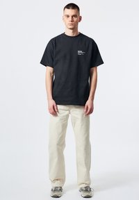 Camiseta de algodón negra con un pequeño gráfico blanco en el lado izquierdo del pecho, combinada con pantalones de corte relajado en color blanco roto y zapatillas grises.