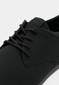 Primer plano de un zapato de cuero negro texturizado con cordones redondos y una etiqueta en la lengüeta con la marca "ALDO."