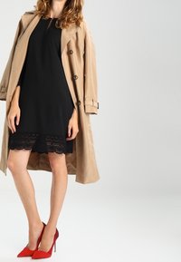 Kvinna klädd i en svart klänning med spetskant, beige trench coat och röda högklackade skor som står framför en enkel ljus bakgrund.