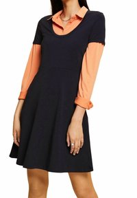 Granatowa sukienka z krótkimi rękawami na pomarańczowej bluzce, z okrągłym dekoltem i sylwetką w kształcie litery A. Gładki materiał i złożone mankiety.